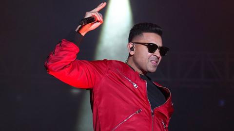 Jay Sean, a man holding a microphone on stage, wearing a red fleece and sunglasses