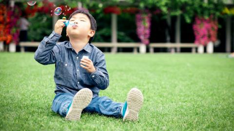 Child sitting on grass blowing bubbles.