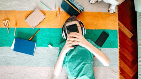A boy lies on a rug with headphones on using a smart phone.
