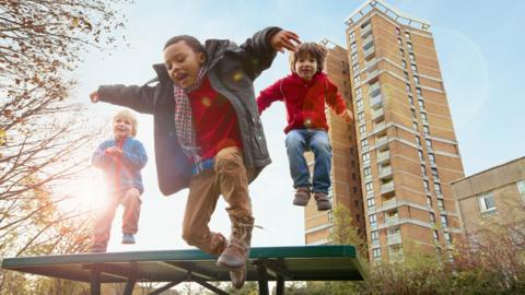 three kids jumping off a platform with highrise flats int he background