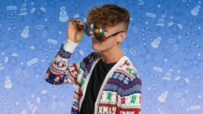 Joel from Blue Peter is wearing a Christmas cardigan and Christmas tree sunglasses. He is tilting the sunglasses down and peering at the camera.