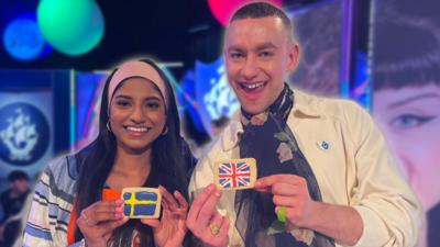 Blue Peter presenter Shini is smiling whilst holding an iced biscuit with the Swedish flag on it. Next to her stands Olly Alexander, who holds another biscuit but with the Union Jack flag on it.