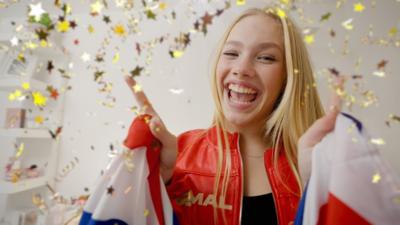 Freya Skye in red jacket smiling. Girl with blonde hair and gold confetti around her. She is holding a union jack flag.