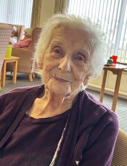 A 100-year-old woman in the lounge of a care home. She is wearing a purple top and cardigan and is smiling, She has short, grey hair