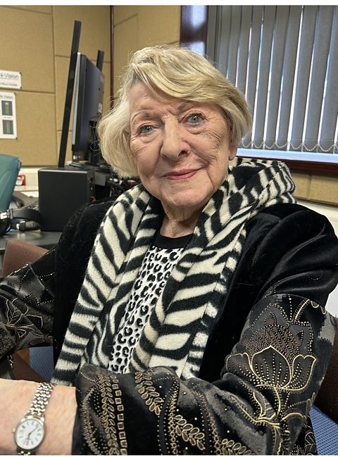 Image shows a 100-year-old woman with ash blonde hair wearing an animal print top and scarf, a black coat and silver watch. She is sitting in a recording studio