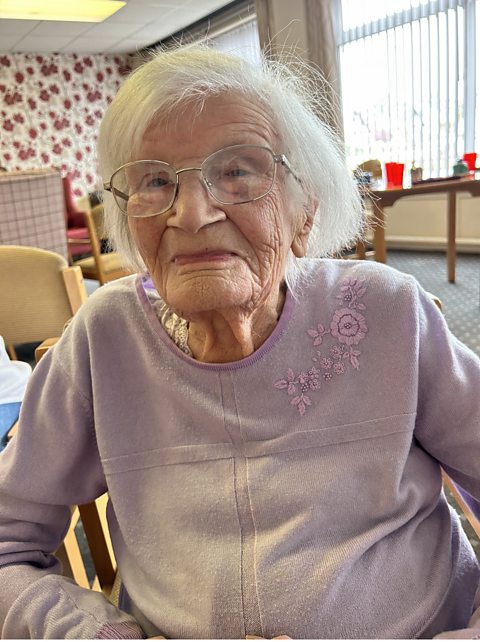 A 103-year-old woman sat in the lounge of her care home. She is wearing a lilac top and glasses and has white hair