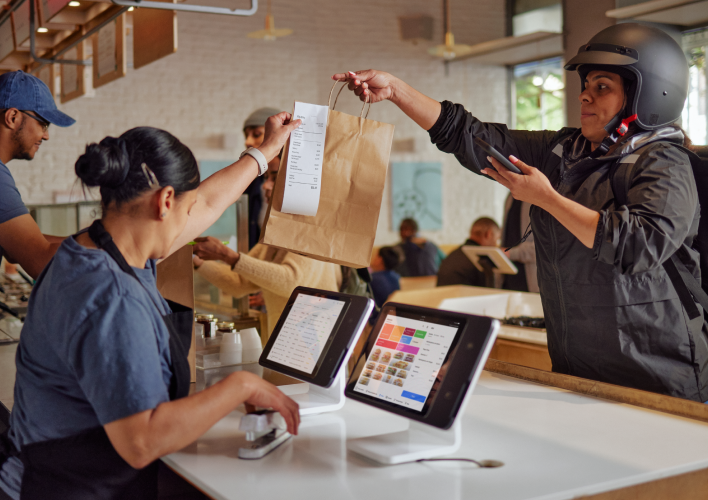 Two restaurant workers work on the register while handing a take-out bag to a delivery person to the right.