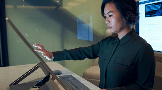 Stranski profil ženske v temnem krilu v pisarni med brskanjem oz. pri delu v napravi Microsoft Surface Studio.
