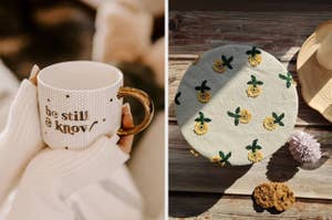 A hand holds a mug with the text "be still & know." Next to it, a round fabric cover with flower embroidery is placed on a wooden surface with a cookie and flower