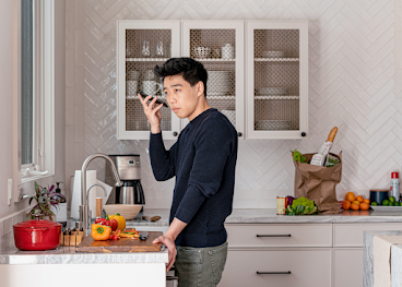 An Asian-American man stands at a kitchen counter holding his Android phone, listening to a screen reader verbalising a recipe.