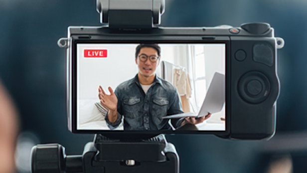 An Asian man records himself and is seen wearing glasses and holding a laptop in the camera's viewfinder