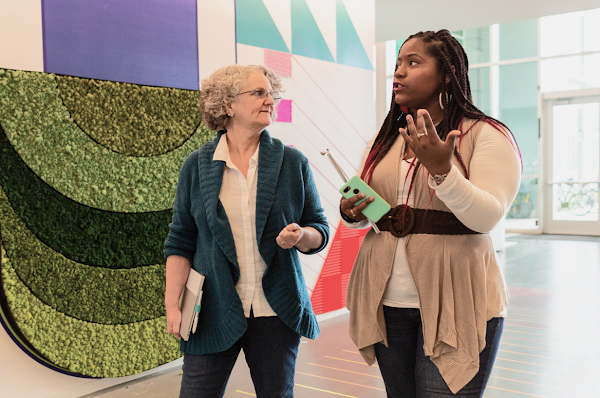 A Black woman with long braids and an older White woman with short gray hair walk engage in conversation as they walk through a hallway with geometric patterns on the wall