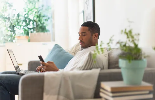 man sitting on couch with a laptop in his lap and a mobile phone in his hand