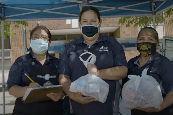 Three female food workers wearing face masks holding a clipboard and bag of food