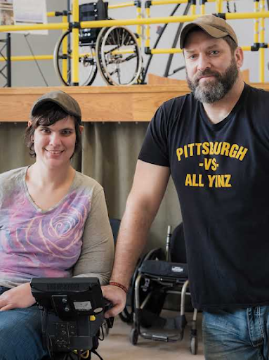 A woman in a motorized wheelchair sits smiling next to a standing man with a cap and beard