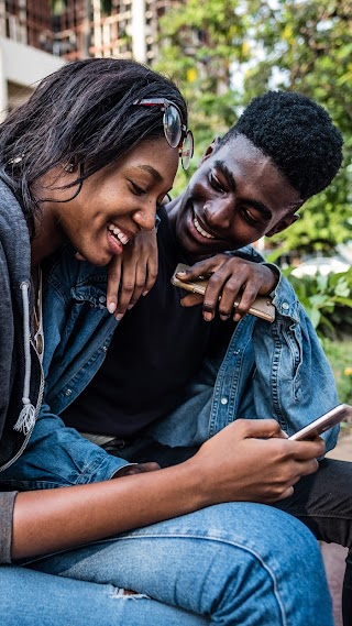 Two young people sitting on a bench, smiling and looking at their phones.