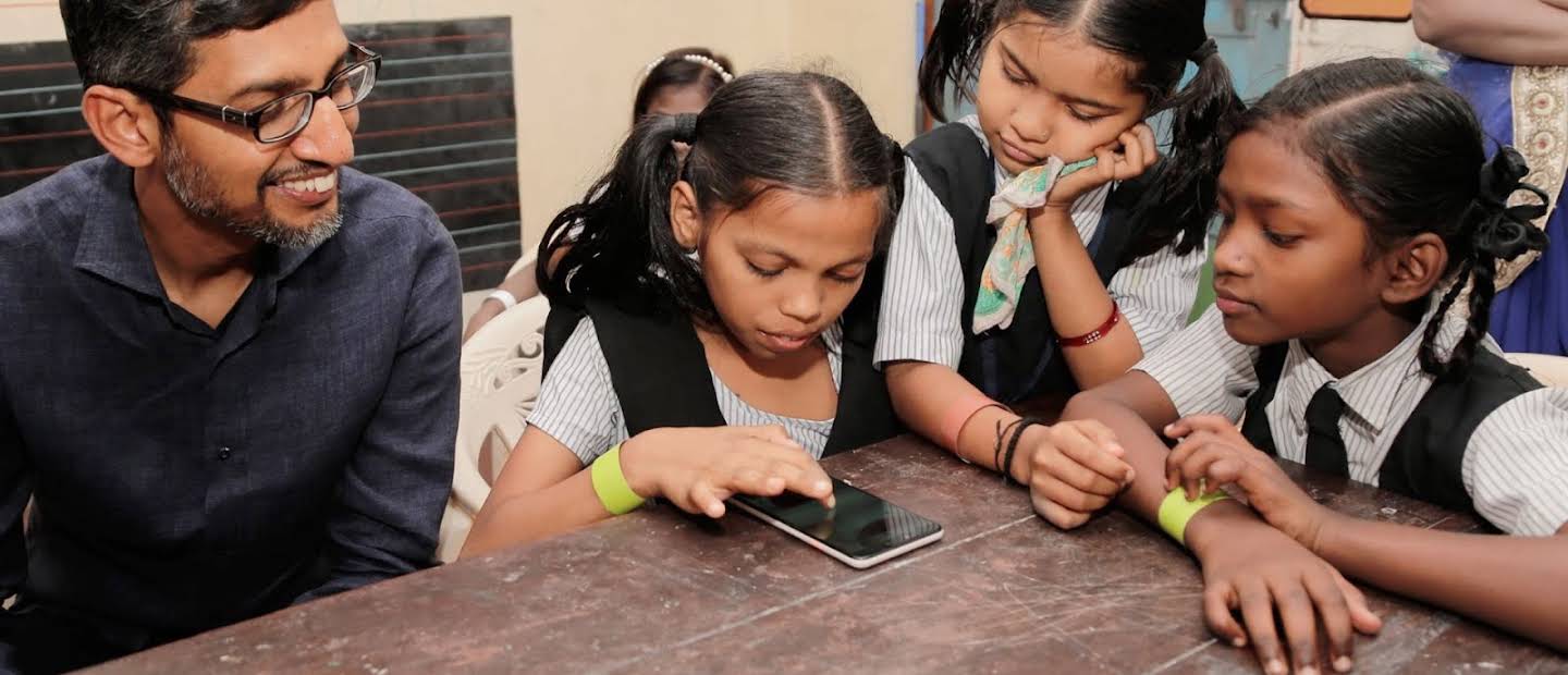 Sundar Pichai i trzy dziewczynki w mundurkach szkolnych skupieni na smartfonie.