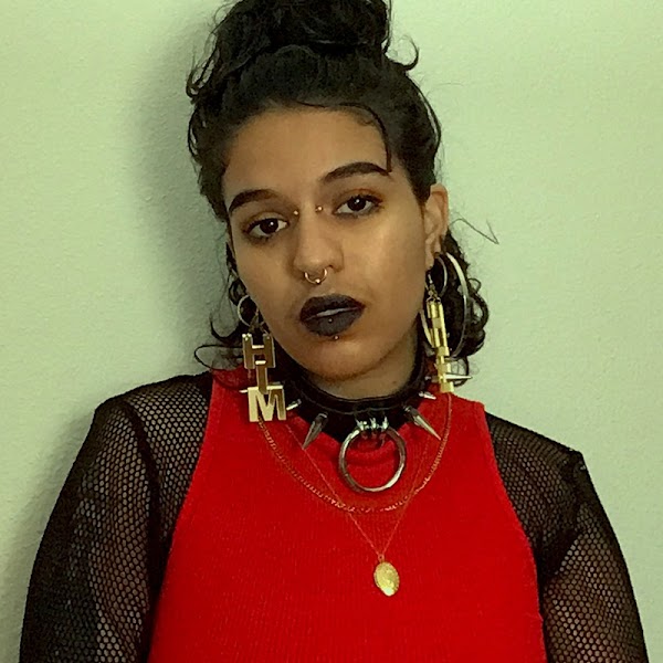 Portrait of artist wearing a red and black top with black lipstick in front of a green background.