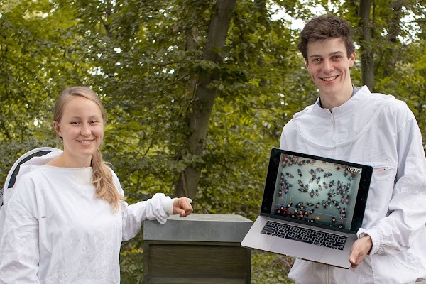Katharina and Frederic standing in front of a beehive, with footage from the camera on a computer