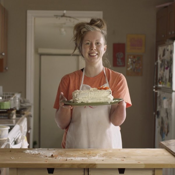 Alexis Hillyard (she/her), a woman with limb difference, stands in a kitchen and holds a cake