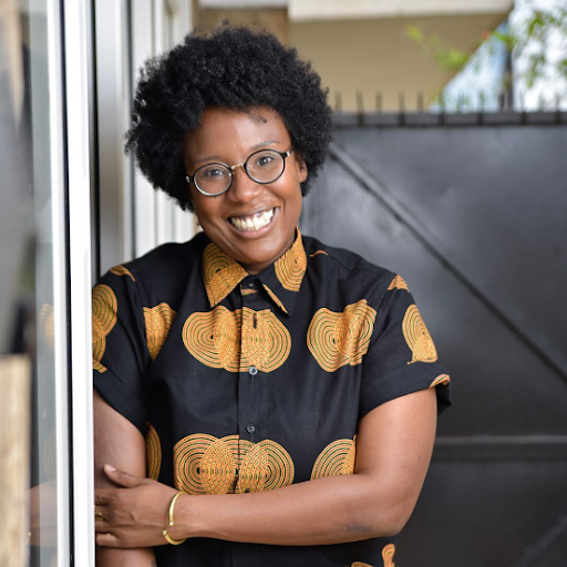 A Senegalese-American woman wearing glasses and a black and orange patterned shirt crosses one arm over the other and smiles