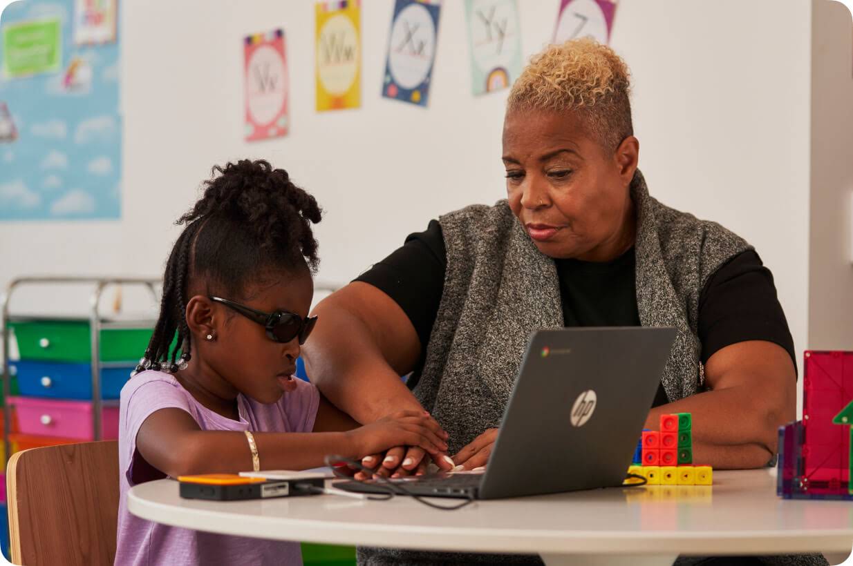 Una alumna joven con discapacidad visual utiliza un Chromebook con la ayuda de un lector de braille mientras la profesora, sentada a su lado, guía su mano.