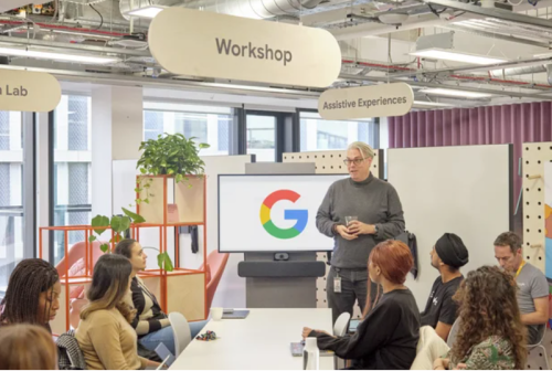 A group of eight people sit around a conference table in the Accessibility Discovery Centre, listening to a man next to a screen with the Google logo on it.