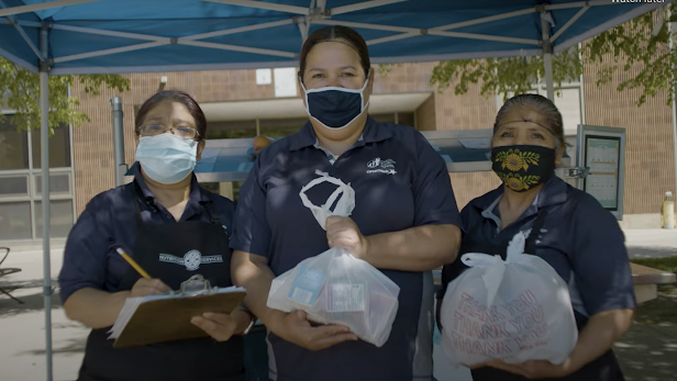 Three female food workers wearing face masks holding a clipboard and bag of food