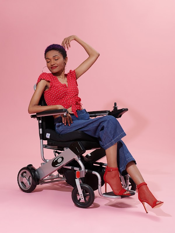 Model in a wheelchair with a red top and red boots in front of a pink background.