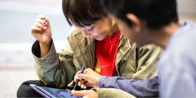 There are two children playing on a tablet. One of them is teaching the other how to use the device.