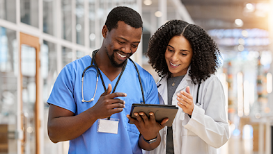 A Black man in blue scrubs wearing a stethoscope reviewing data on a tablet with a Black, female doctor in white scrubs in a healthcare center.