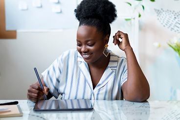Smiling black woman wearing a striped blouse and diamond shaped earrings in a well lit office space working on a tablet with a stylus.