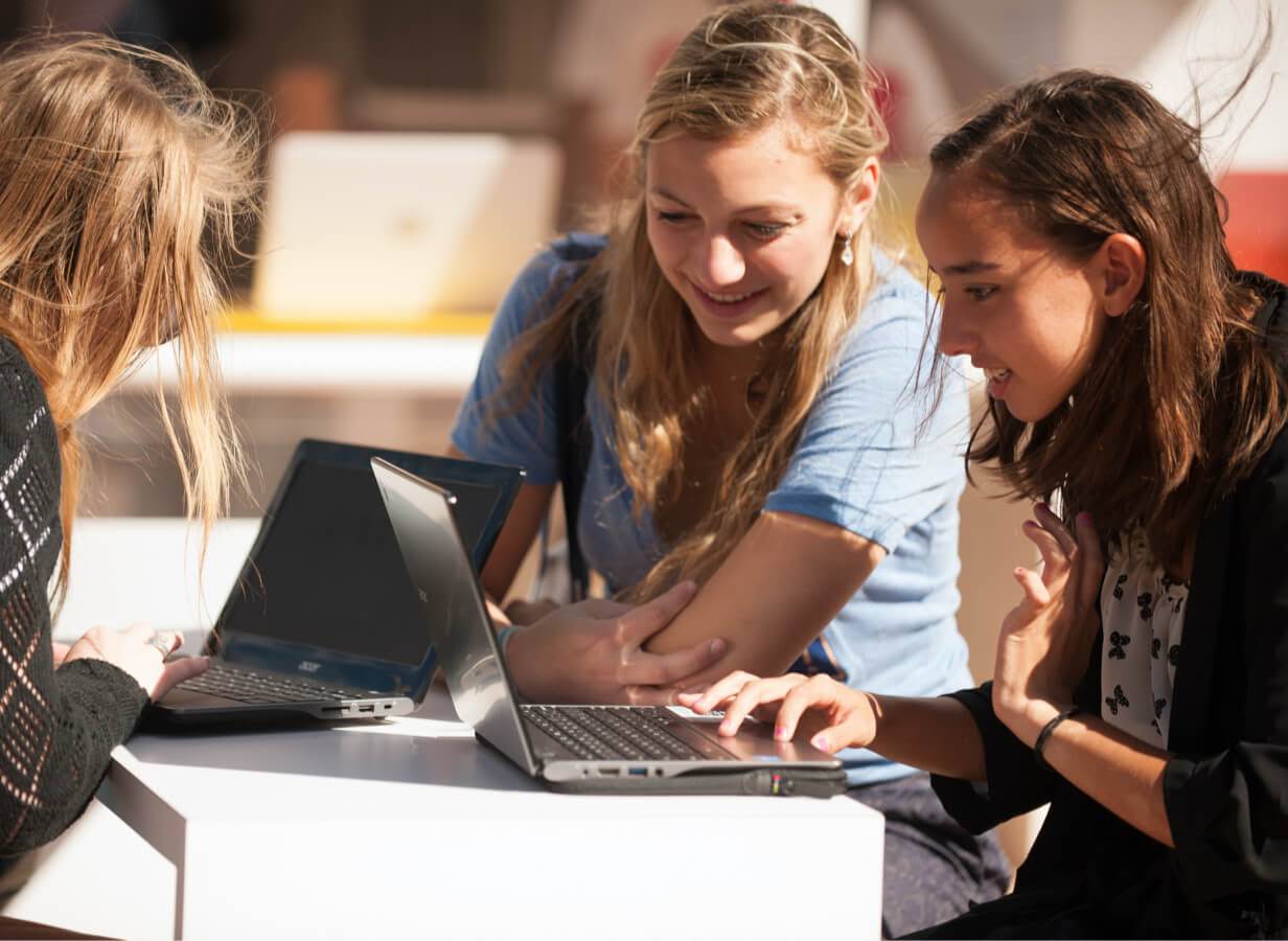 Tiga siswa duduk di meja di luar ruangan, sedang menggunakan Chromebook mereka