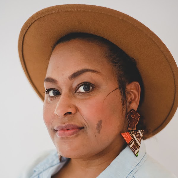 Portrait of artist in a caramel colored wool hat with orange and brown geometric shaped earrings.