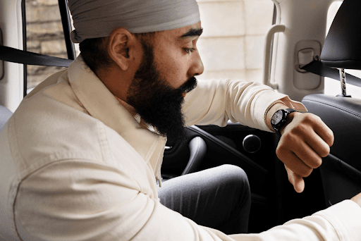A man is in the back seat of a car with his left arm raised closer to his face as he is looking at a notification on his smartwatch. He is leaning forwards and holding the back of the driver's seat, appearing to be on the go and in a hurry.