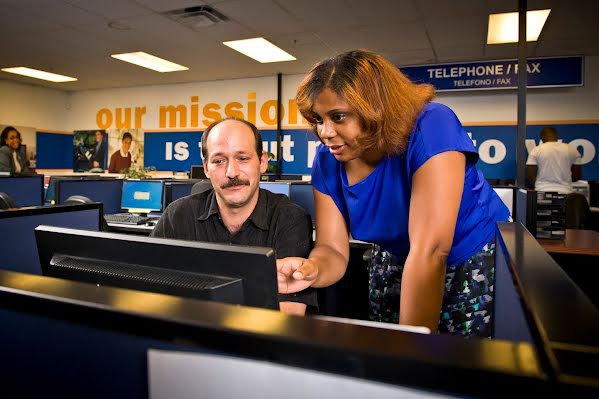 A man sitting at a computer, as a woman stands pointing at the screen delivering instructions.