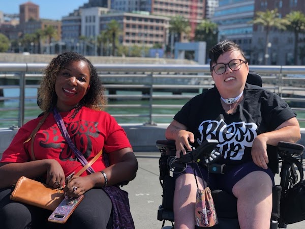 Stacey Park Milbern and Andraéa LaVant smile at the camera while seated in power chairs at the front of a ferry.