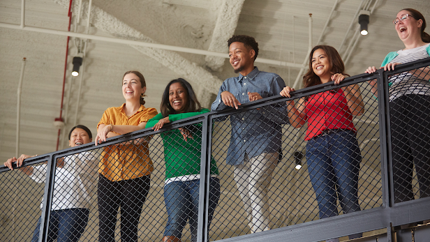 A group of happy Google employees in brightly colored shirts look down from the railing of a workplace stairwell