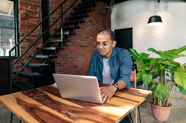 A man with glasses sits at a high top wooden table while working on his laptop.