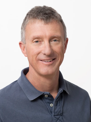 A smiling man with short grey hair and blue eyes wears a dark blue collared shirt