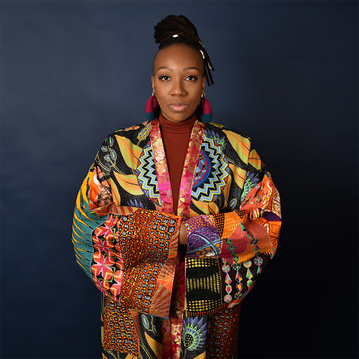 Portrait of artist with their arms crossed wearing a multi-colored kimono style jacket with geometric shapes in front of a black background.