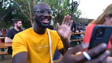 Black man wearing glasses and a yellow shirt waves at the phone he’s holding up