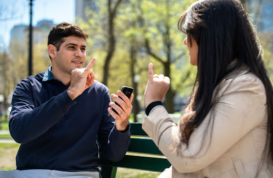 A man and woman sitting on a park bench, speaking sign language with the assistance of a mobile phone.