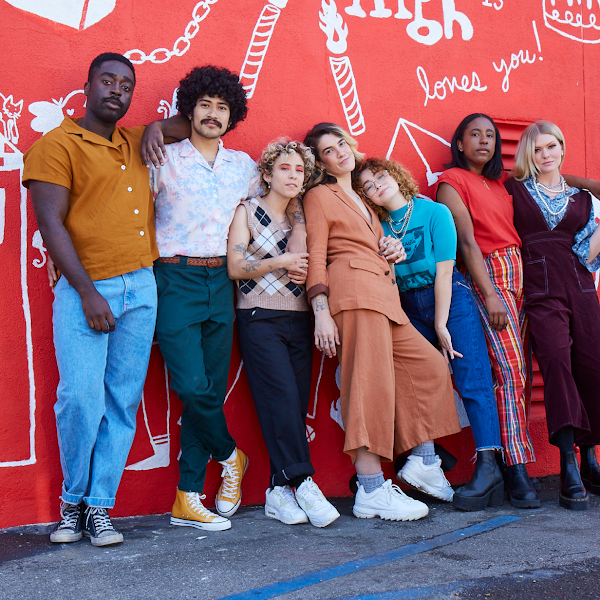 A group of seven people wearing colorful outfits stand outside in front of a red wall with white illustrations
