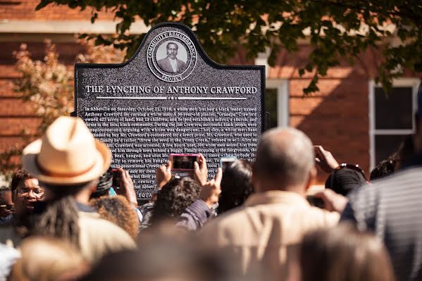 A group of people outdoors, taking photos of memorial plaque that reads "The Lynching of Anthony Crawford."