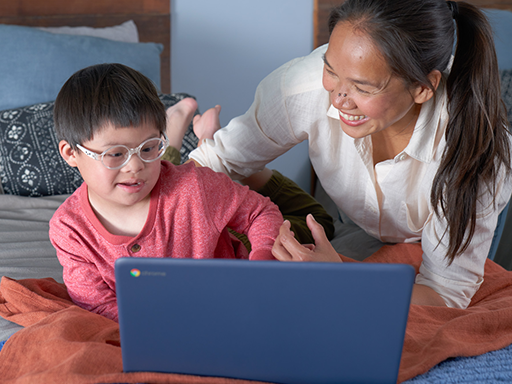 A smiling woman looks over the shoulder of a child with learning differences as he uses his computer