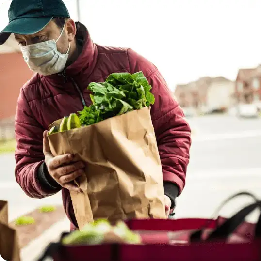 A 412 Food Rescue employee wearing a green cap and a disposable face mask holds a paper grocery bag containing lettuce, bananas, and other foodstuff; before him, a graphic reads, “50% cut down of reporting time”
