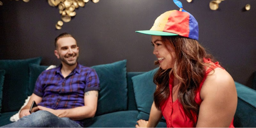 A woman with long hair is seated on a couch wearing a red blouse and a hat with Google’s yellow, red, blue, and green colors. There is a man sitting next to her also smiling.