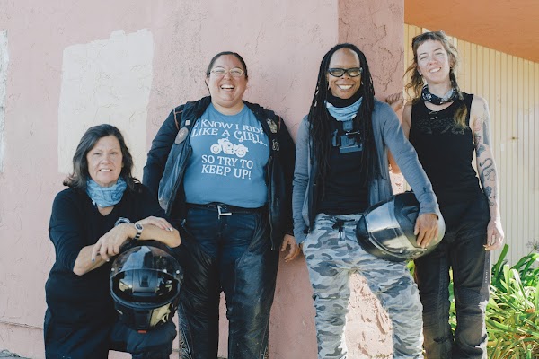 Four women wearing motorcycle gear face the camera smiling