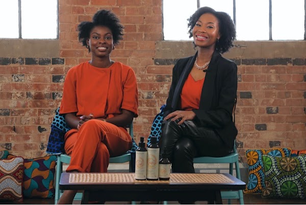 Rachael and Joycelyn, founders of Afrocenchix, smile sitting side by side. Both have short, textured hair. Rachael wears an orange outfit, Joycelyn wears a black suit and orange top.